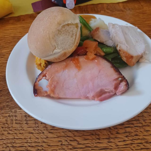 White plate piled with a slice of ham, turkey, a bread roll, green beans, and sweet yams.