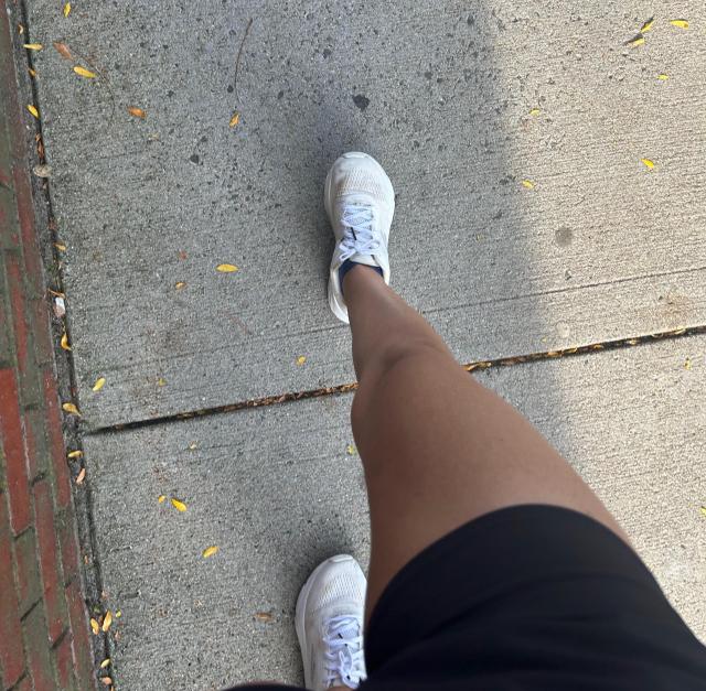 A girl's tan leg wearing black running shorts and white running sneakers, mid stride on a concrete sidewalk.