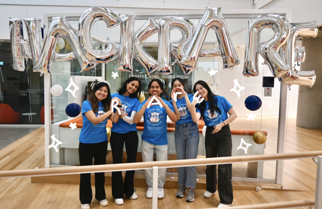 A group of five students wearing blue shirts in front of silver balloons that read "HACK RARE." They are making symbols with their hands to spell out "RARE."