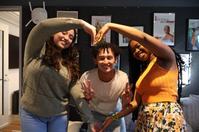 This is a picture of three smiling individuals posing together by forming a heart shape with their arms. The person on the left wears a green sweater and has long, wavy hair. The person in the middle, wearing a white T-shirt, flashes peace signs with both hands, while the person on the right, dressed in a bright orange top and floral skirt, completes the heart shape with a big smile. The background features framed posters and a cozy room ambiance.