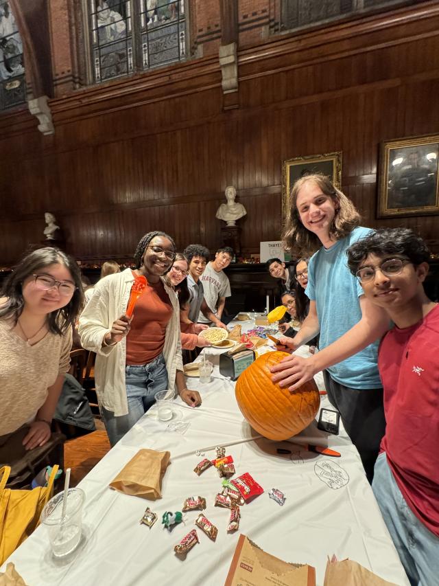 Students holding pumpkin-carving tools stand around a table in Annenberg Hall. On the table sits a pumpkin and several pieces of candy.