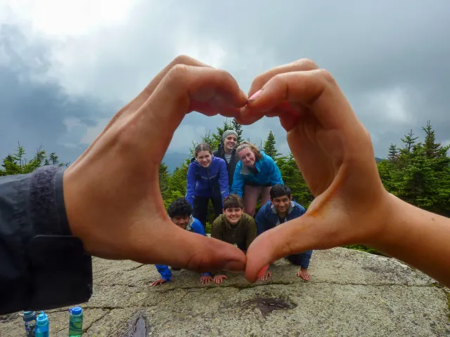 A group of six first-year students on a FOP trip form a human pyramid atop a mountain summit, framed by the hands of their FOP leaders, which form a heart shape.