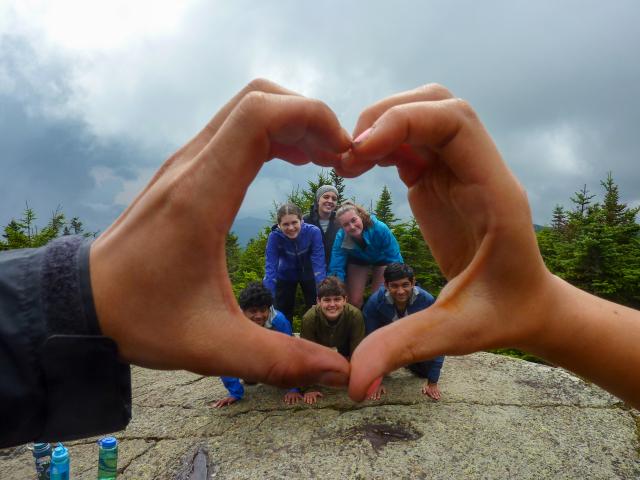 A group of six first-year students on a FOP trip form a human pyramid atop a mountain summit, framed by the hands of their FOP leaders, which form a heart shape.