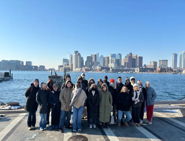 A group of people in winter clothing standing on a waterfront with a city skyline in the background under a clear blue sky.