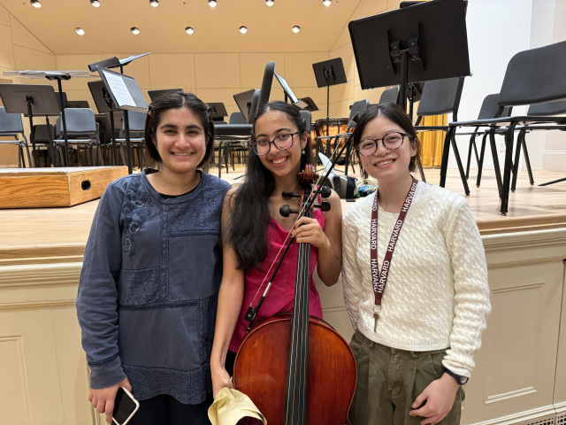 Meena holding a cello at the end of her concert, smiling and standing beside two of her roommates who came to celebrate with her.