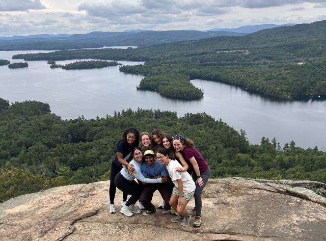 Lizbeth and seven peers posing on top of a mountain in New Hampshire, which a large lake in the background.