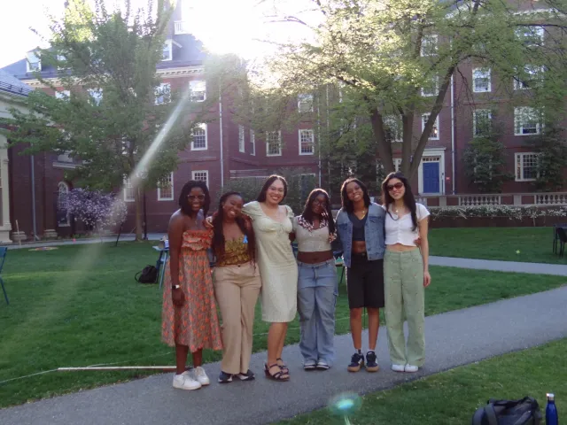 A group of six friends stand close together and smile for the camera on a sunny day in a grassy courtyard at Harvard. They are dressed in casual spring outfits, with sunlight streaming through the trees behind them and red-brick buildings in the background.