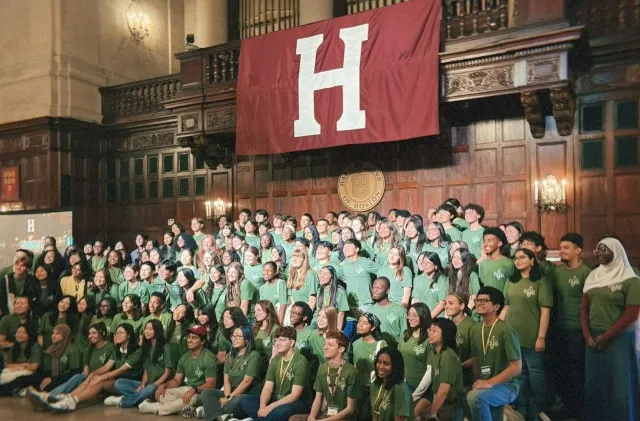 A large group of diverse students wearing matching green t-shirts posing for a group photo inside a grand, wood-paneled hall. They are gathered beneath a large crimson banner displaying a white letter "H" and a circular Harvard Club of Boston emblem.