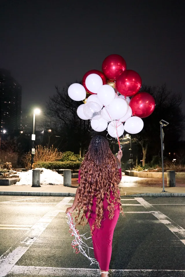 Photo of girl in red dress holding red and white balloons while walking across the street