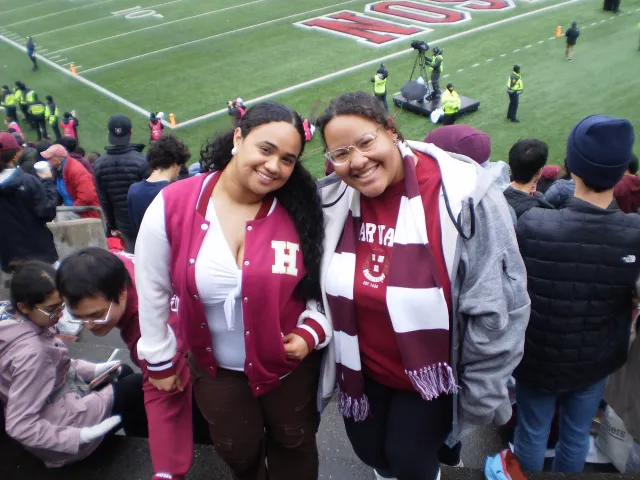Two best friends smiling at the camera in the stands at Harvard vs Yale game.