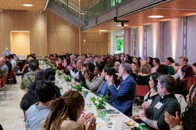 Many students, faculty, and staff sit at several long tables, eating and talking
