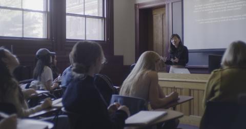 A teacher lectures from the front of a class room. Students sit at desks and take notes