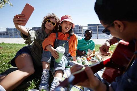 4 students sitting on a lawn in front of the river haning out, playing music and taking pictures