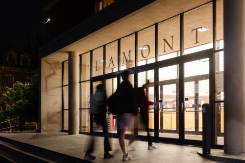 Students entering the front of Lamont Library at night with the glass entrance lit up from the inside.