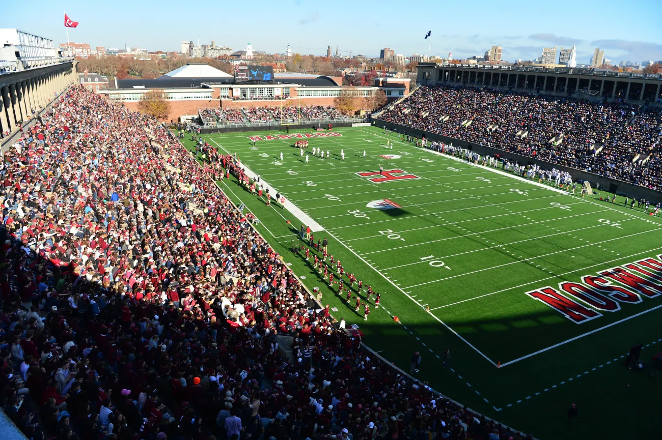 An upward view of Harvard Stadium filled with fans watching the Harvard Crimson football team on the field.