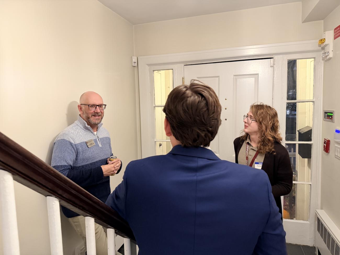 Three people stand chatting by a stairway in a hallway near a white door.