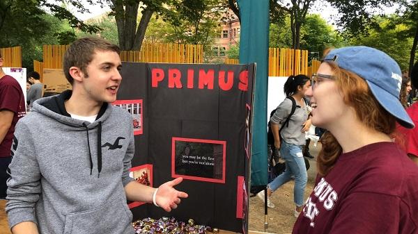 two people talking outside next to a poster that says Primus