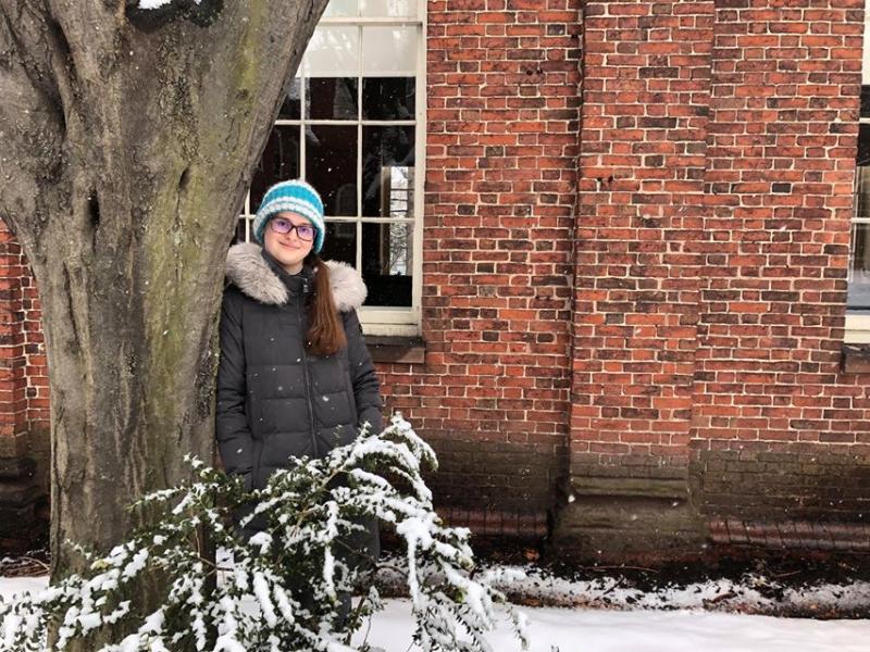 A photo of a girl wearing a gray coat and green beanie, leaning against a tree on a snowy day