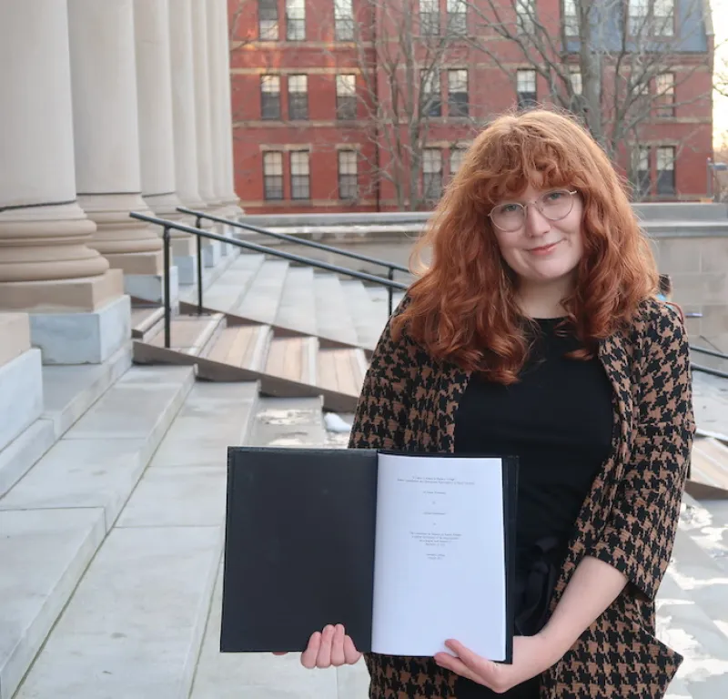 Allison standing on the steps of a large library holding a black thesis binder opened to the title page of her thesis.