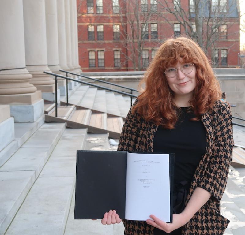 Allison standing on the steps of a large library holding a black thesis binder opened to the title page of her thesis.