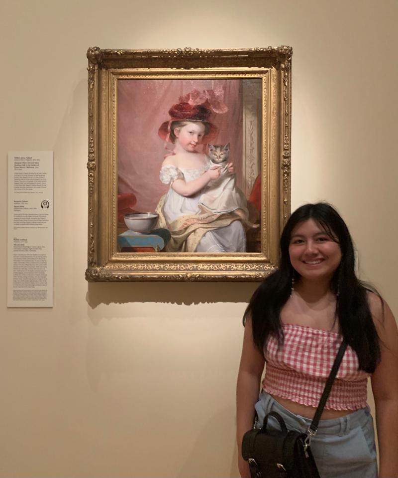 A girl standing in front of an painting of a young girl holding a cat.