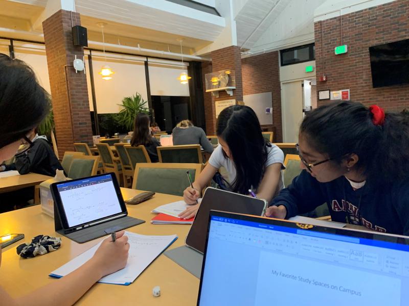 A college dining hall with picturing three girls studying with tablets and laptops.