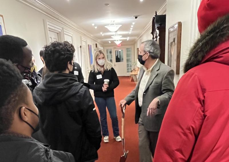 President Bacow, holding a shovel, speaking to students around him in the administrative building.