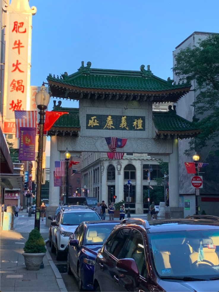 A picture of the Boston Chinatown gate from the sidewalk with parked cars lining the road.