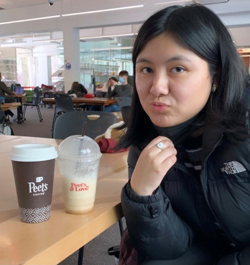 A girl posing next to two coffee drinks sitting down at a table.