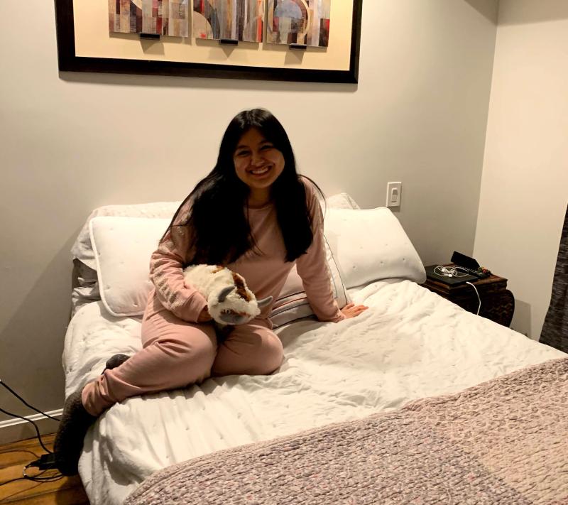 A girl with pink pajamas holding an Appa pillow (from the TV show Avatar) lounging on a bed with white pillows and a pink blanket.