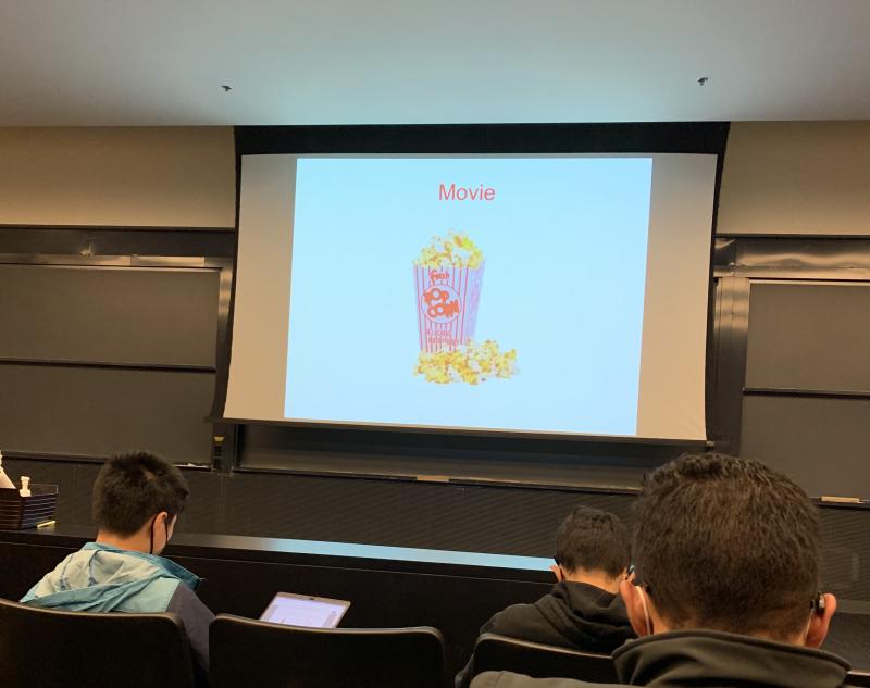 Students sitting in front of projector screen with a picture of popcorn and the word &quot;movie.&quot;