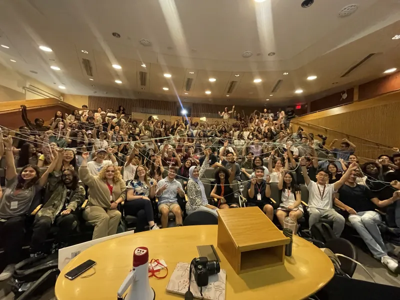 Students sit in an auditorium, and hold sections of the same spool of yarn.