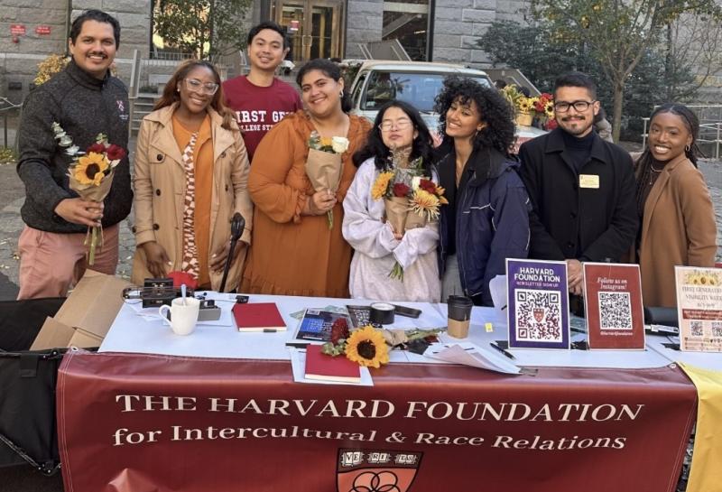 Picture of students holding flowers behind a table with a "Harvard Foundation for Intercultural & Race Relations" banner.
