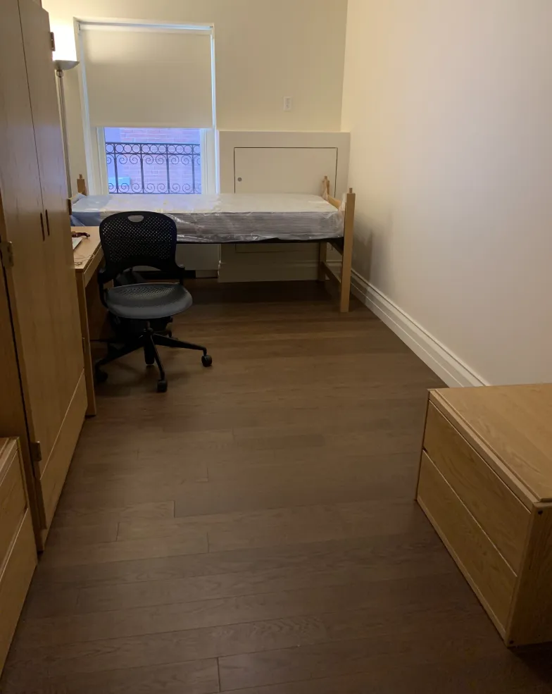 A dorm room with a wardrobe, chair, desk, bed, and dresser.