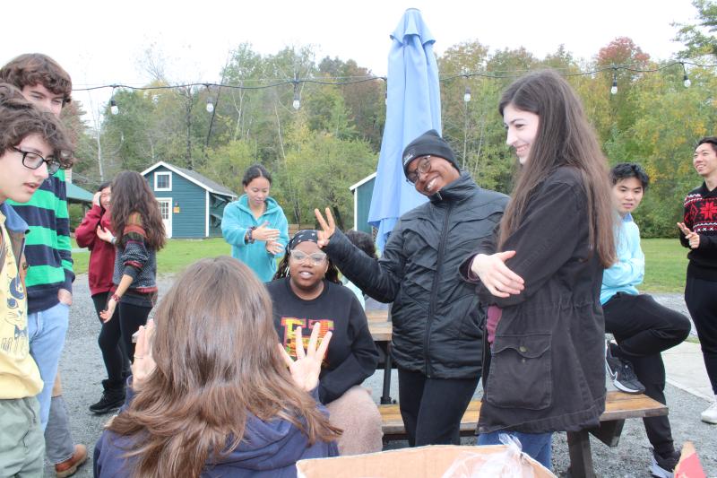 Picture of members of the Harvard-Radcliffe Collegium Musicum chatting around outdoor picnic tables with green and red trees in the background.