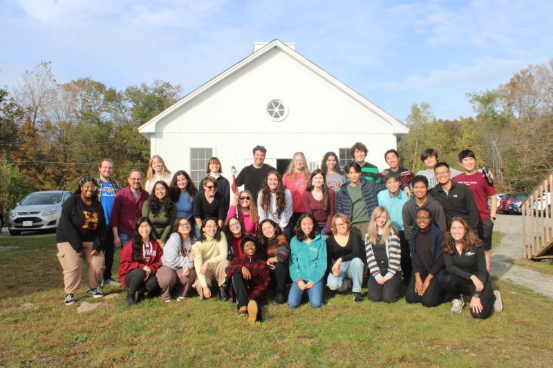 Picture of the Harvard-Radcliffe Collegium Musicum standing in front of a white cabin under blue skies on a green hill.
