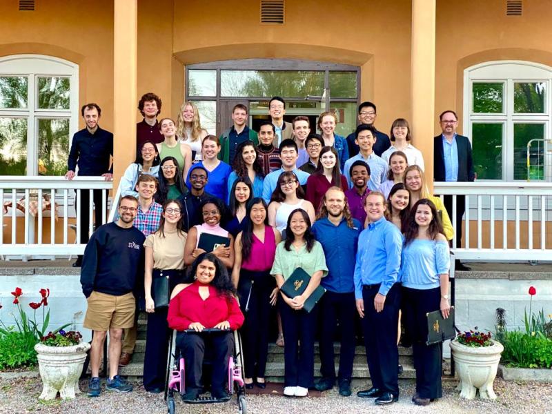 Picture of members of the Harvard-Radcliffe Collegium Musicum standing in front of the entrance of a building with yellow walls and white railing.