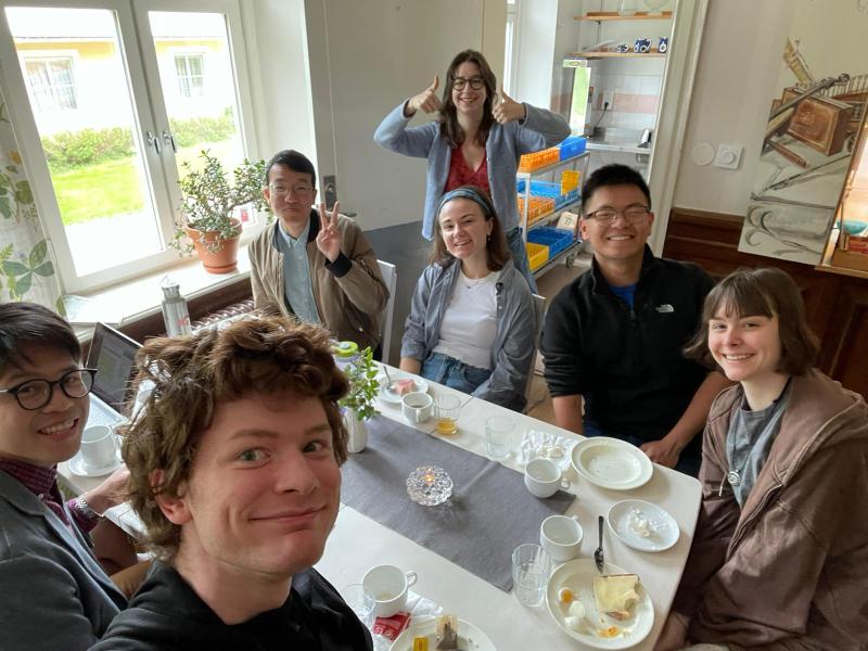 Picture of members of the Harvard-Radcliffe Collegium Musicum sitting around a table with white and gray tablecloth with plates of bread and butter, smiling at the camera.