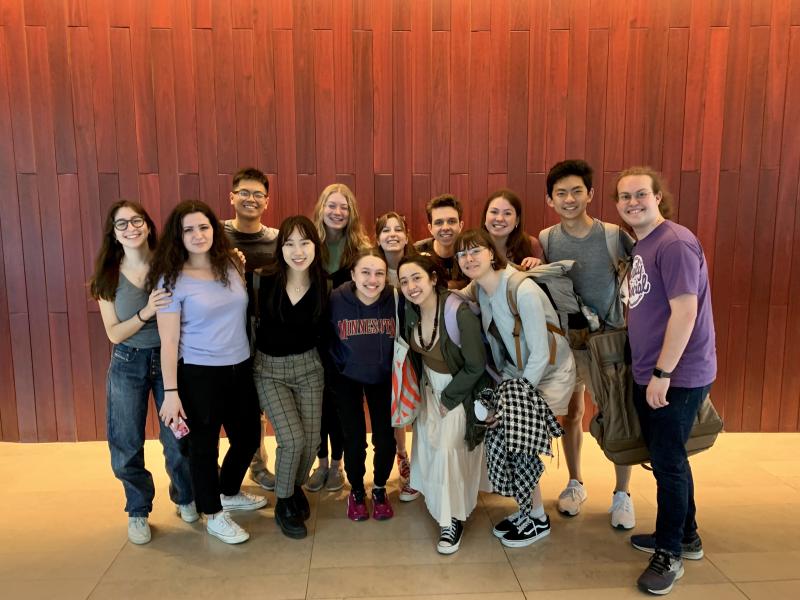 Picture of members of the Harvard-Radcliffe Collegium Musicum standing in front of a maroon wooden background on a yellow-tiled floor.