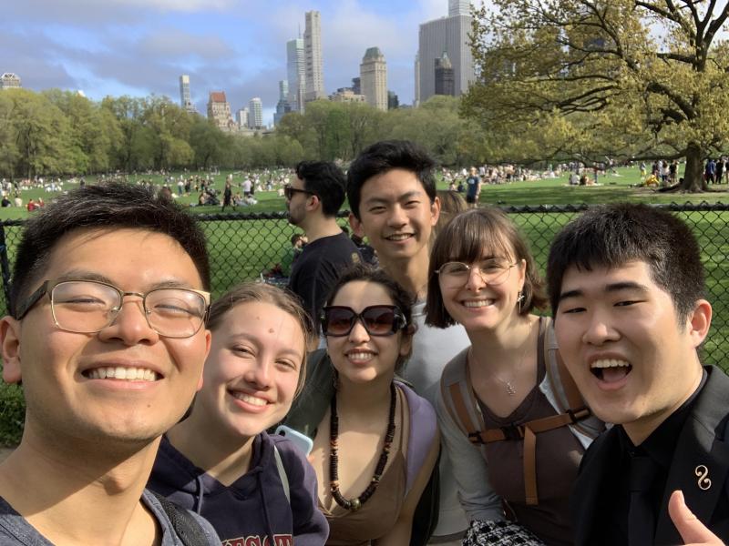 Picture of Raymond and his friends standing in front of a large grassy park lawn with tall buildings and a blue sky in the background.