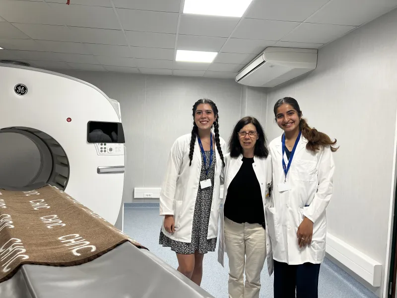Three women in lab coats smiling in front of an MRI machine