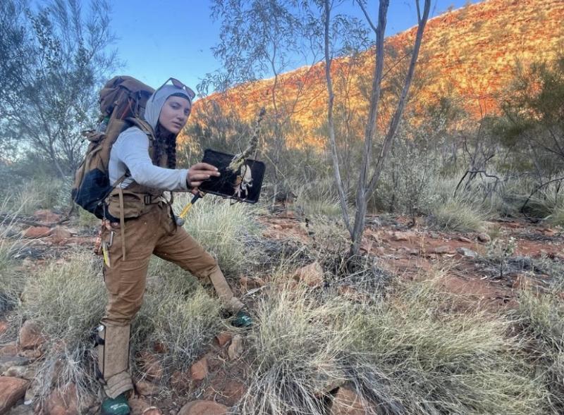 Me with backpacking gear in overalls holding up an ipad so that the Golden Orb spider was visible on its web. 