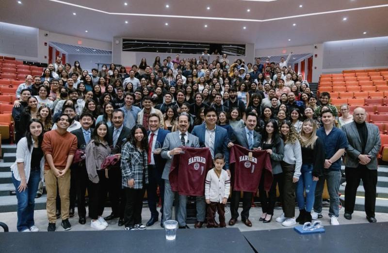 Pictured is a large group of people in a Harvard Science Center Lecture Hall. The large crowd includes band members from Mexican band Los Tigres del Norte.  The band members are holding up a Harvard sweater, and everyone is posing for the group picture, most with a smile on their face.