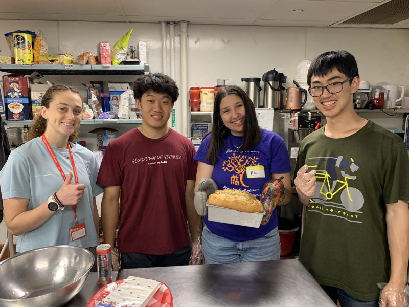 Image of four people standing together in a kitchen, smiling toward the camera, with one holding bread in a bread pan.