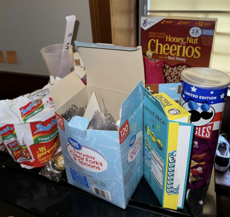 A picture of the top of a black mini fridge, with many snacks like pringles and cereal, as well as plastic utensils