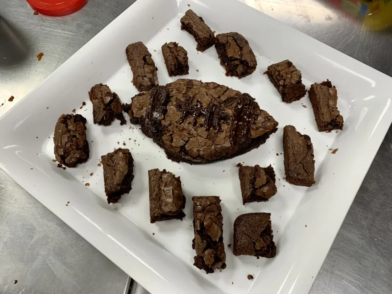 A picture of brownies on a large square, white serving tray with one large brownie in the shape of a football in the middle.
