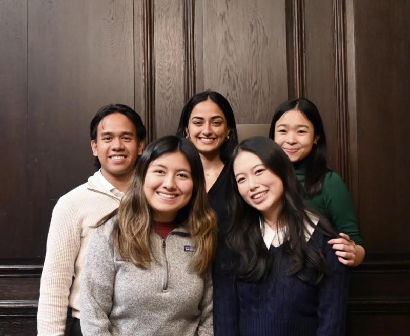 A group of five students standing against a dark brown wooden wall. Three students in the back, left to right, Marcky, Krisha, and Beverly; two students crouching in the front, from left to right, Kathleen and Janny.