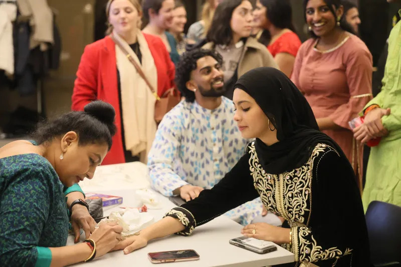 A mehndi artist applies intricate designs using henna as students wait in line for their turn.