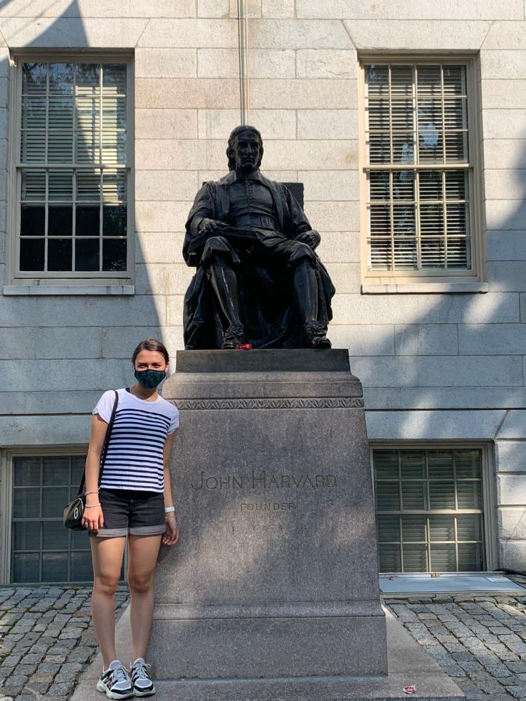 A picture of Ana in front of the John Harvard Statue right before she started her first year at Harvard. it was a warm and sunny day, and she is wearing a face mask (picture was take in August of 2021). 