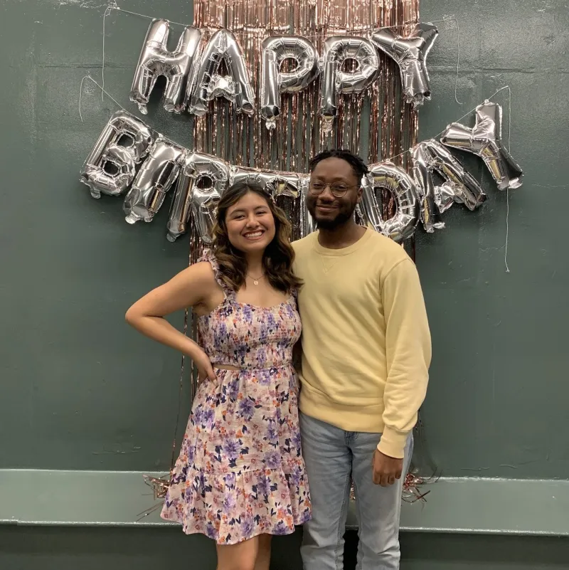 A girl and boy standing together and smiling, with the balloon letters "Happy Birthday" behind them.